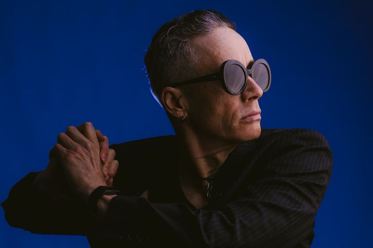 Australian performer iOTA wearing oversized round glasses and a dark suit jacket, posing in profile against a blue studio background.